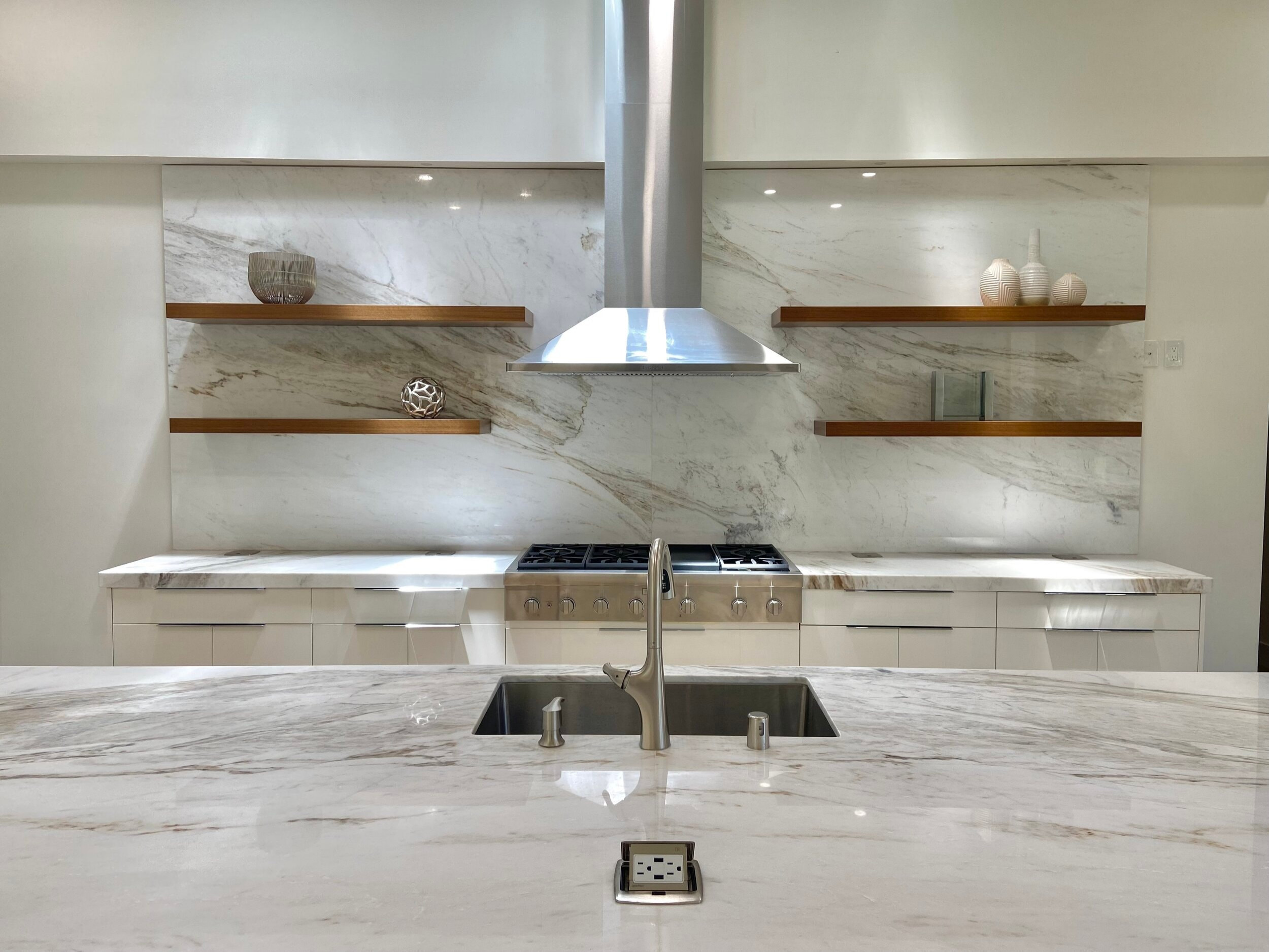 Kitchen island sink facing the range, stainless hood and veined stone backsplash